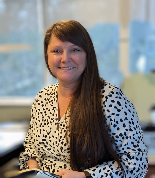 Woman with long dark hair smiles, wearing a black top and colorful necklace; indoors.