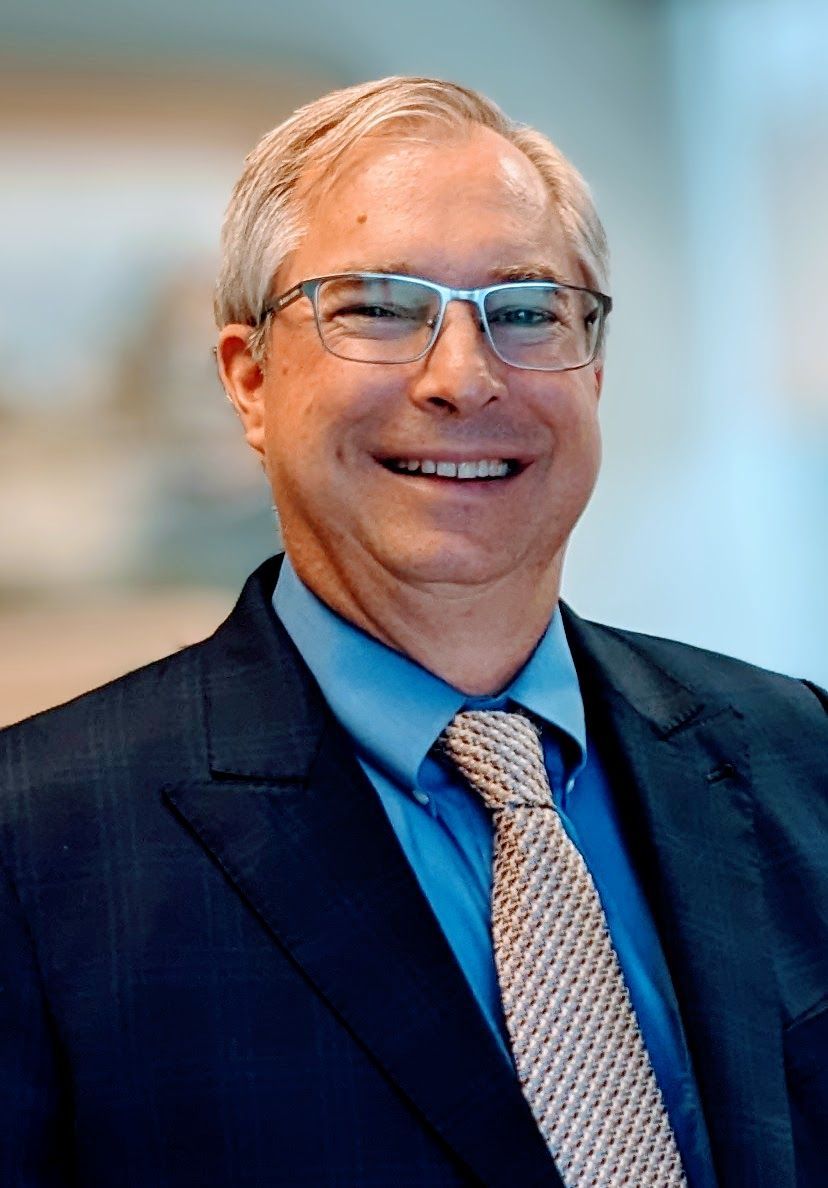 Man with glasses smiles, wearing a blue striped shirt and patterned tie, outside.