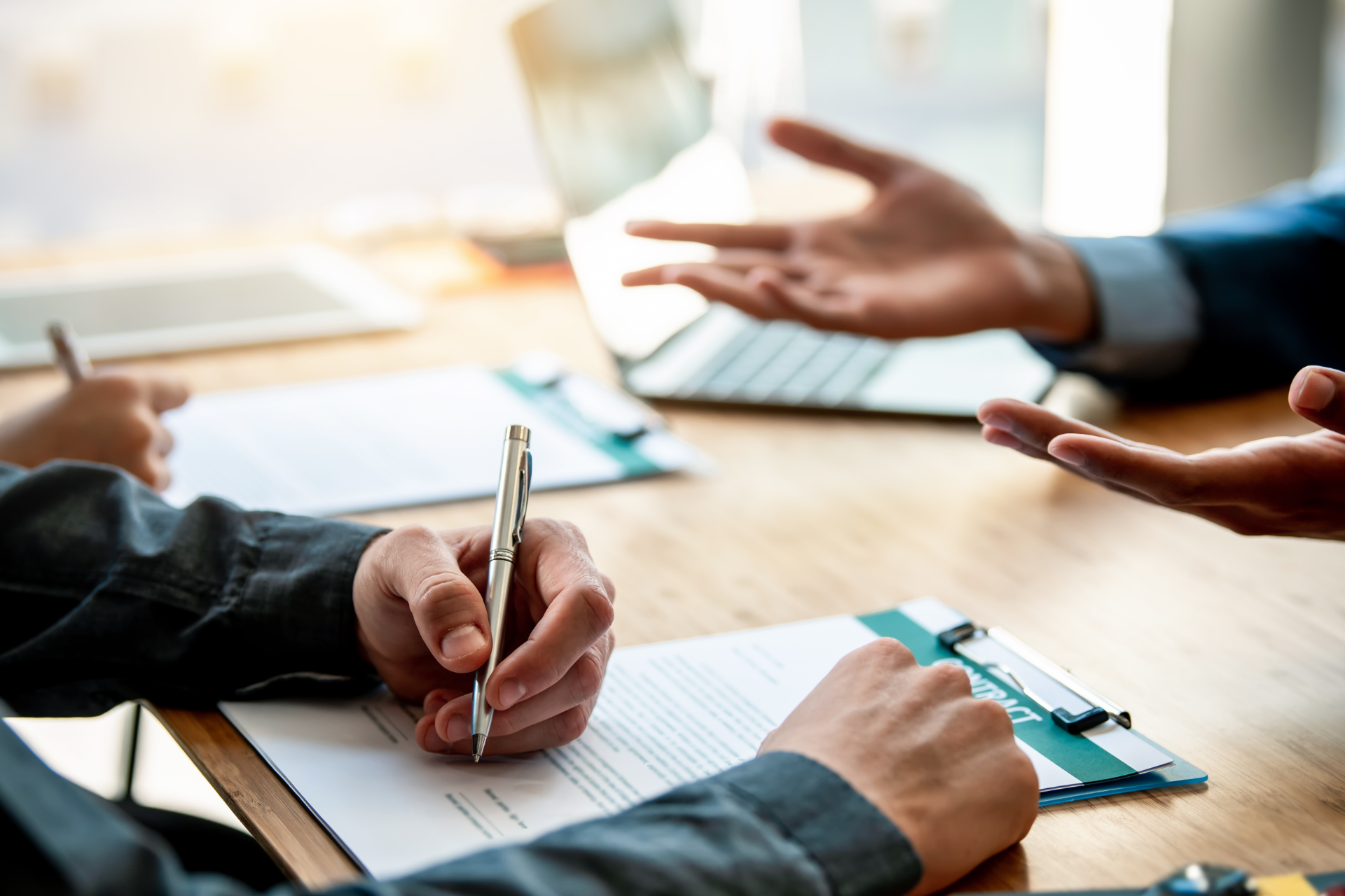 Hands of a person signing documents at a table; others present. Office setting with laptop and tablet.