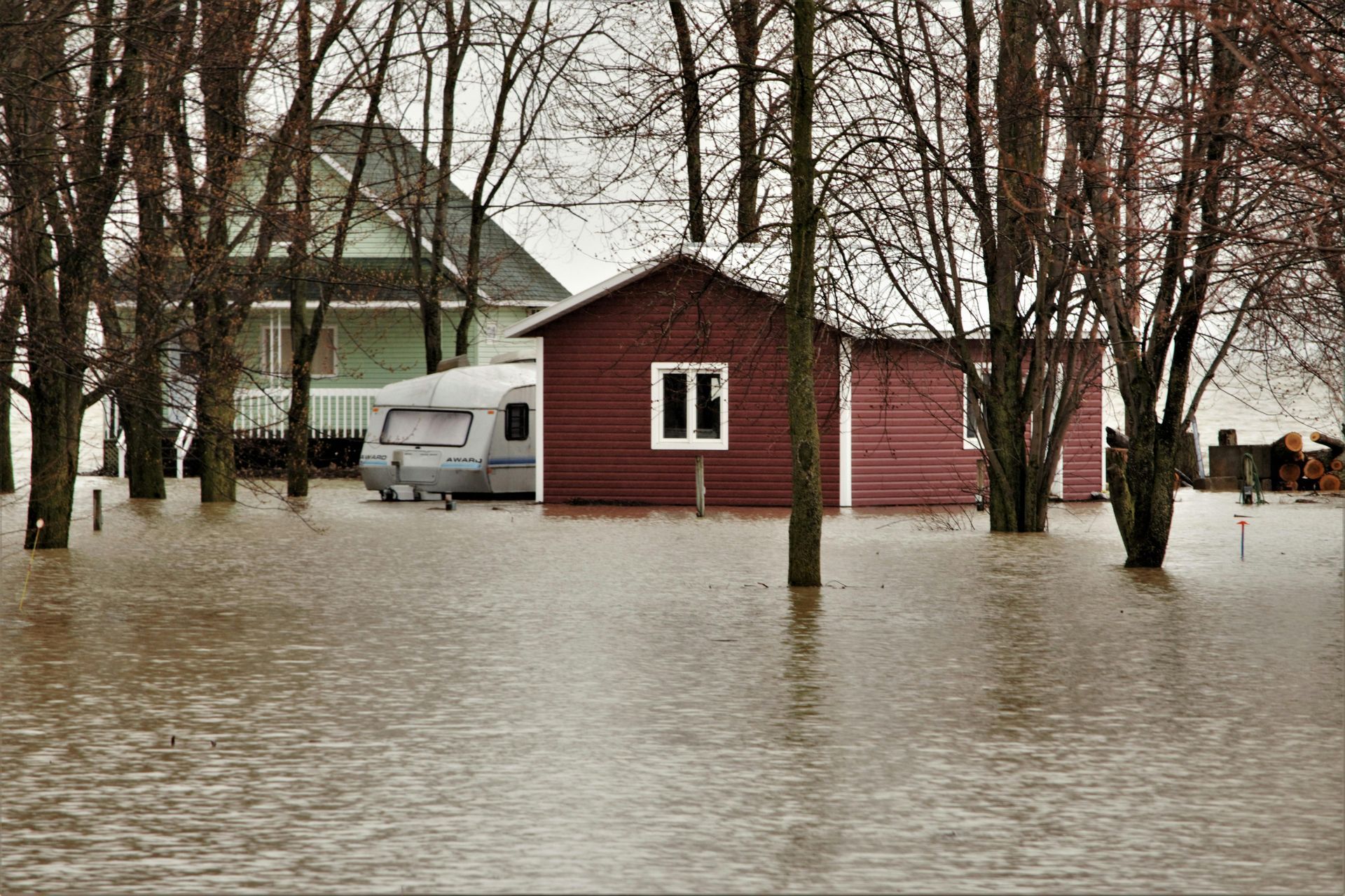 Flooded rural scene: brown water surrounds a red building, trees, and a camper in a cloudy, outdoor setting.