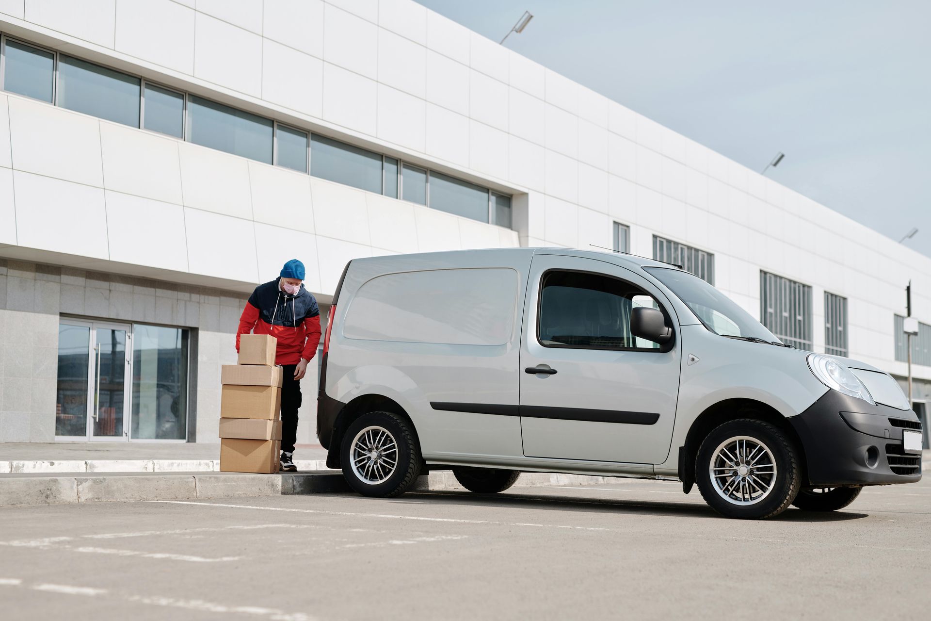 Delivery person loading boxes into a silver cargo van in front of a building.