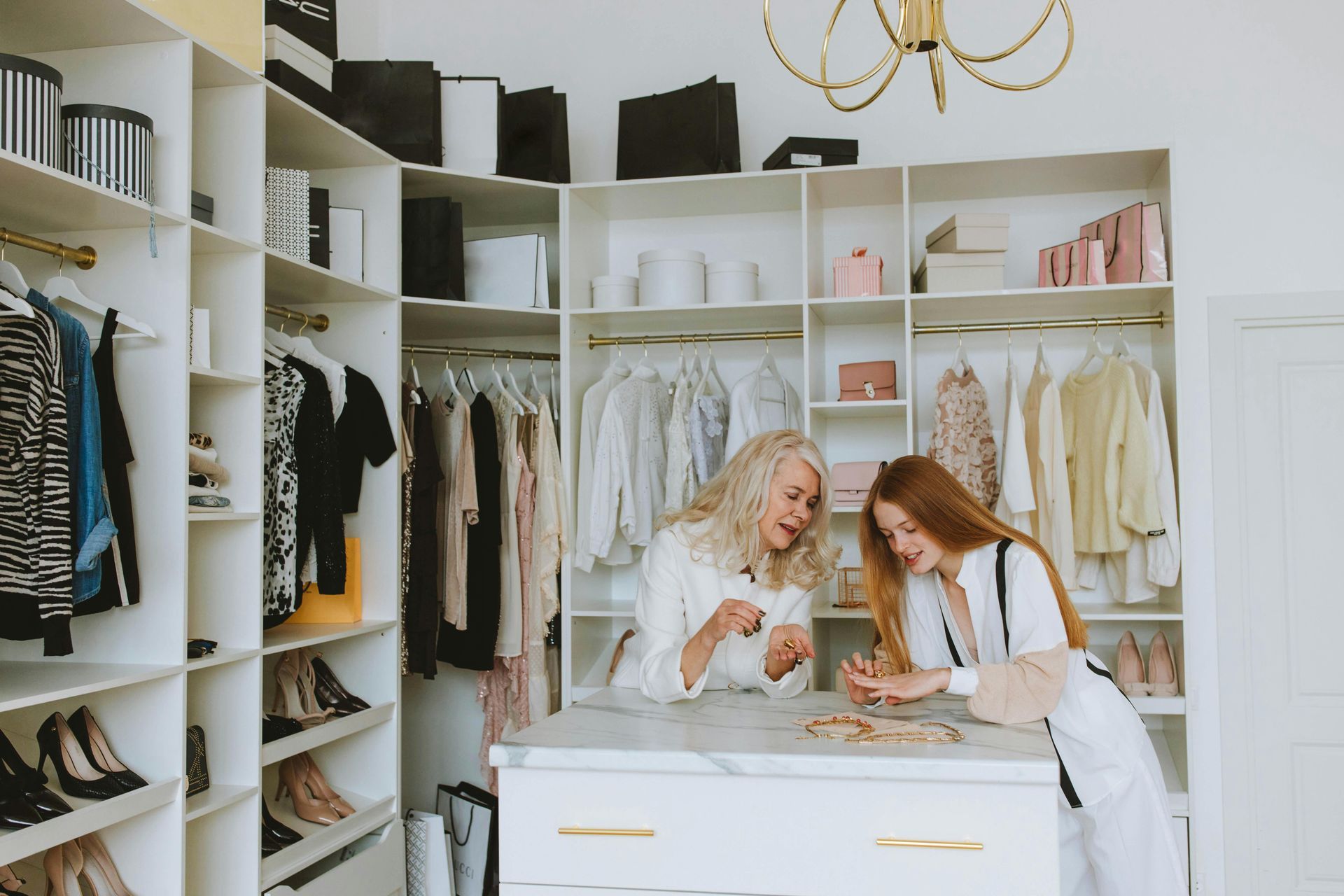 Two women in a white closet, examining jewelry on a marble island, surrounded by clothes and shoes.