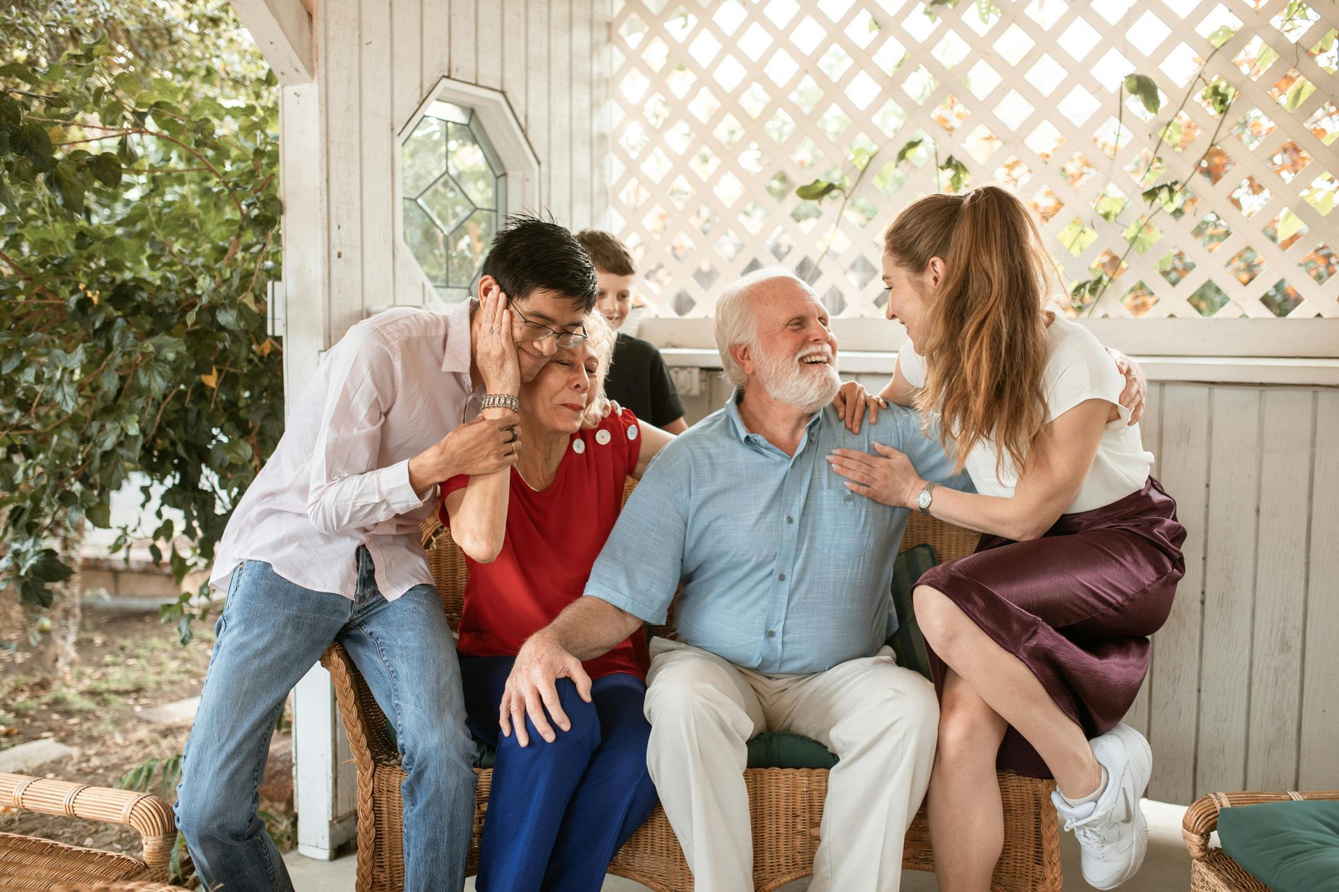 Family gathered on a porch, laughing and embracing. Elderly couple, young adults, casual attire.