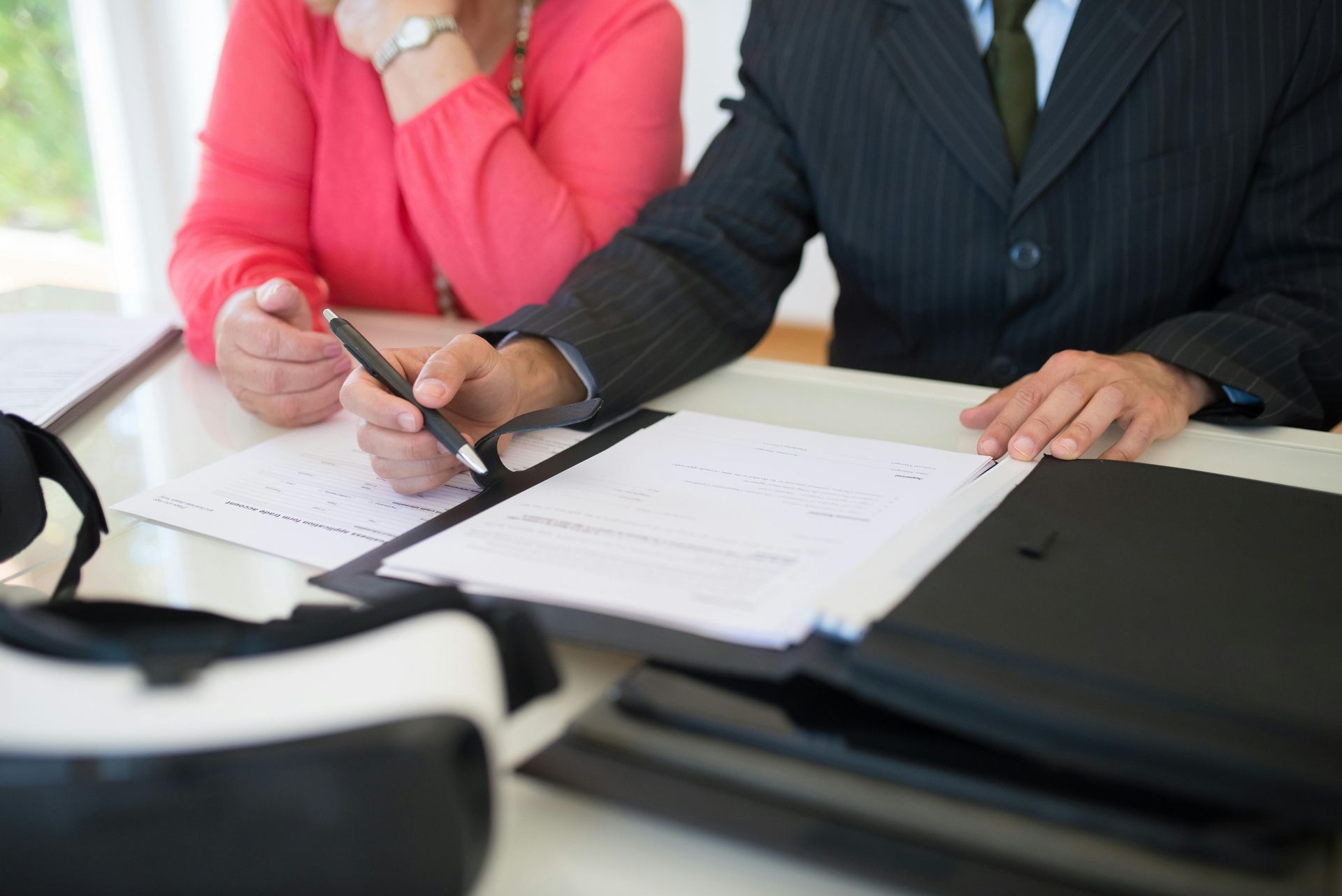 Man in suit signing documents with a woman in pink looking on; VR headset on table.