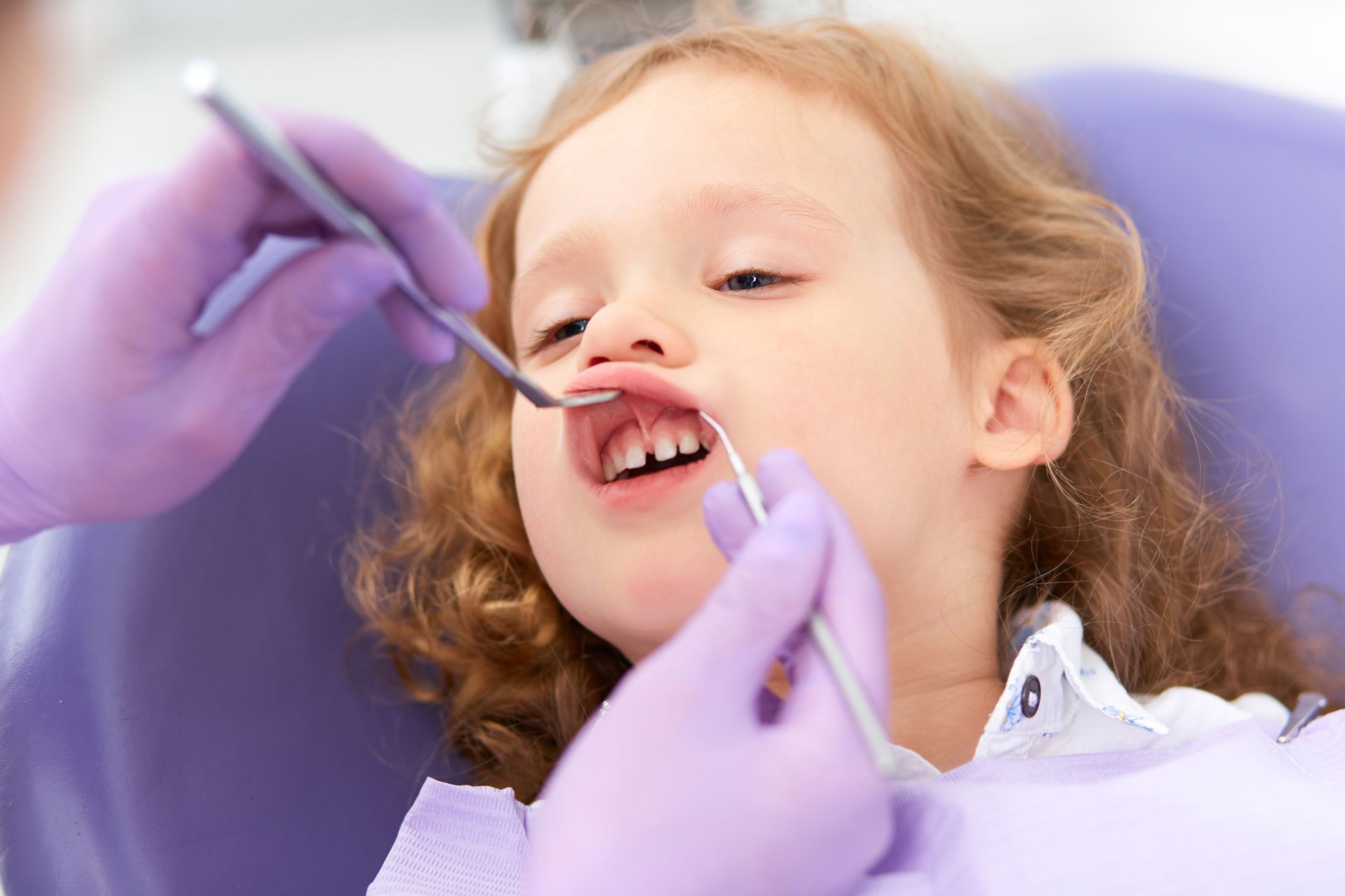 Pediatric dentist with blue gloves checks under the limps of a young and blond kid, inside a dentist’s office.