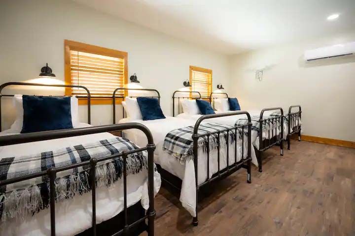 Four beds in a room with white bedding, navy pillows, and plaid blankets; wooden floor and trim.