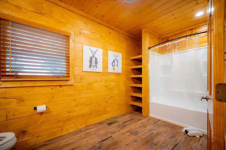 Wooden bathroom with shower, toilet, and shelves, two animal art prints, and a window with blinds.