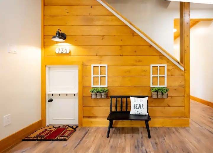 Playhouse built under stairs with a small door, windows, bench, and wooden paneling.