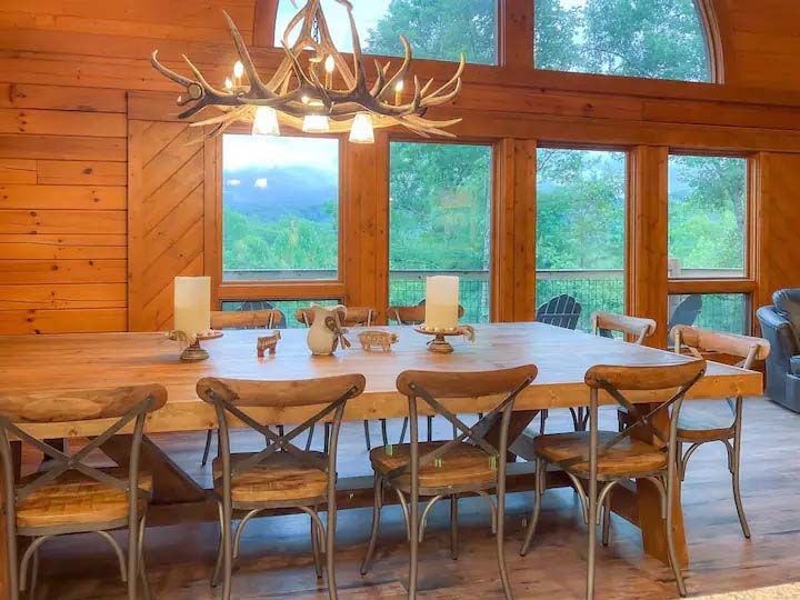Dining room with wooden table and chairs, antler chandelier, and large windows overlooking a green landscape.