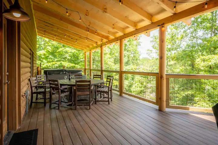 Wooden deck with table and chairs, overlooking a lush green forest, string lights overhead.