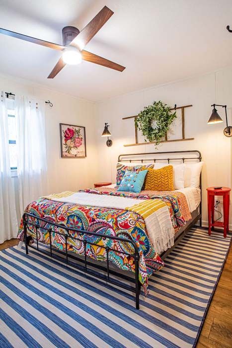 Bedroom with a wrought iron bed, colorful quilt, blue striped rug, and a ceiling fan.