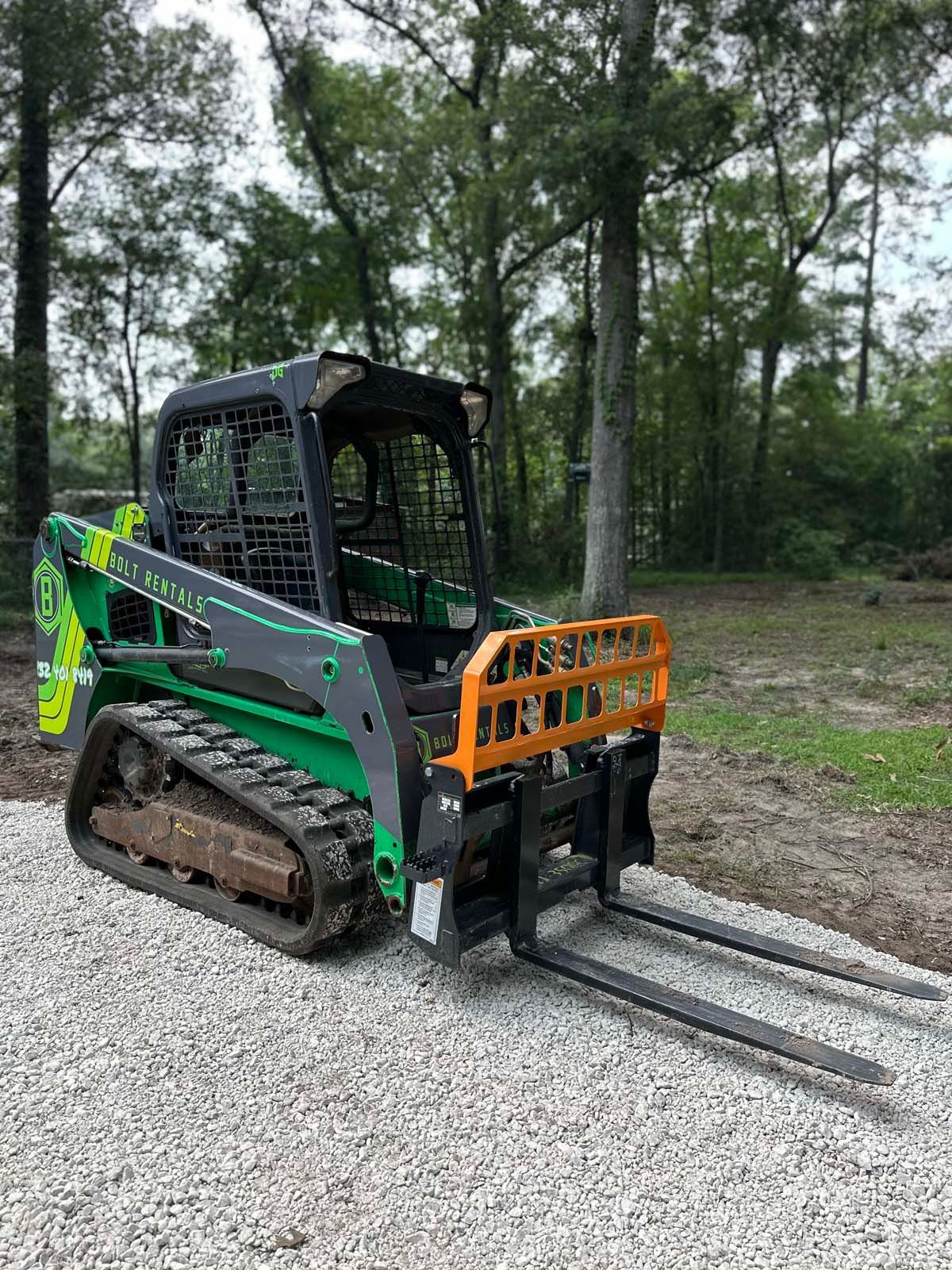 A green skid steer with forks attached to it is parked in a gravel lot.