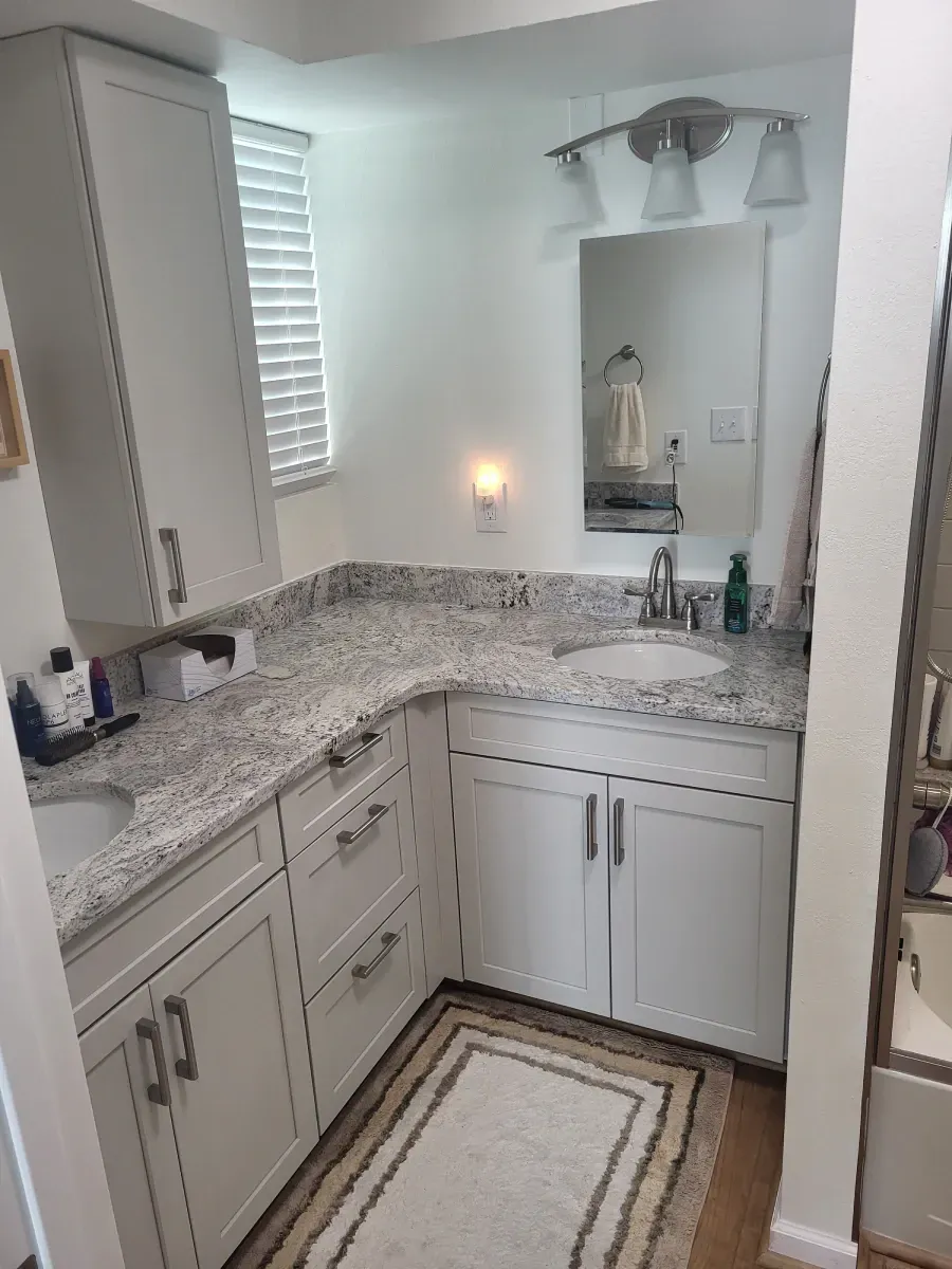 Small white bathroom with a granite countertop, sink, mirror, and cabinet. There is a window with blinds and a rug on the floor.