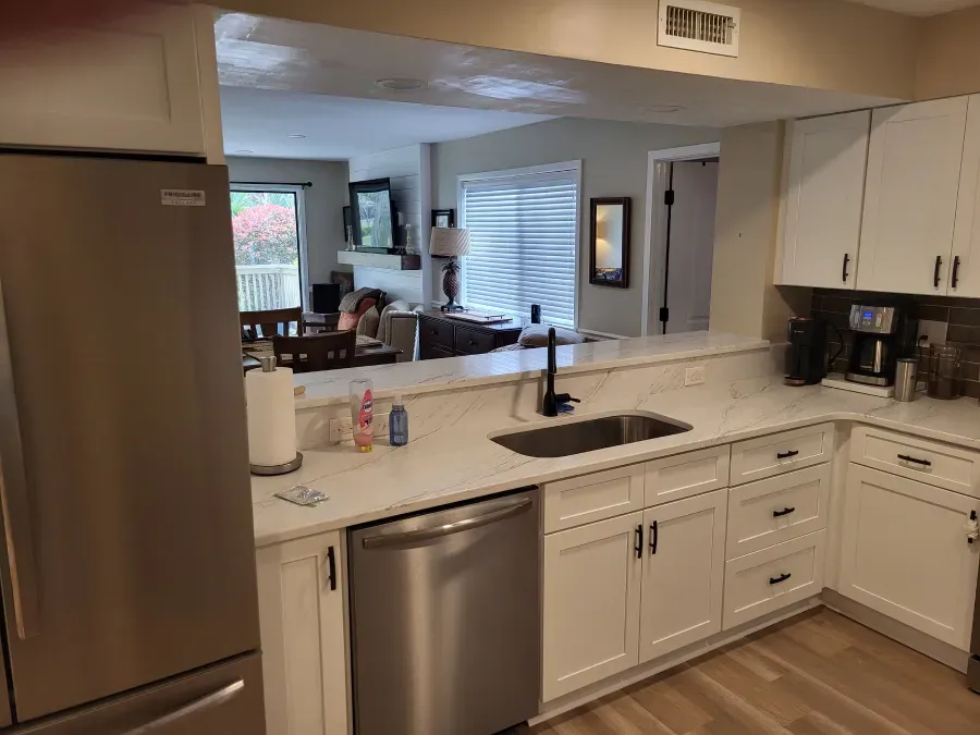 A remodeled kitchen with white cabinets and countertops. A stainless steel dishwasher and refrigerator are present. The kitchen opens to a living area.