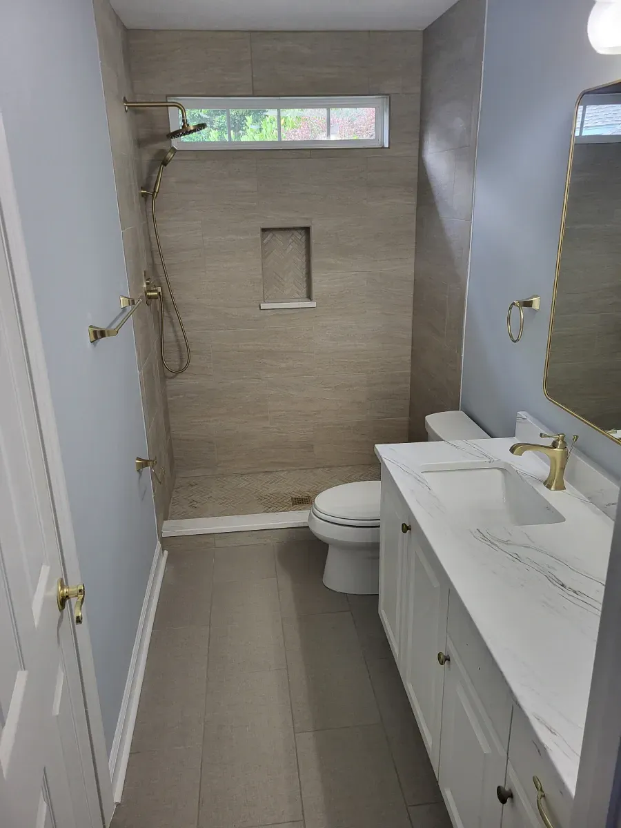 A newly renovated bathroom featuring a walk-in shower, vanity with gold fixtures, and a toilet, with gray and white tones.