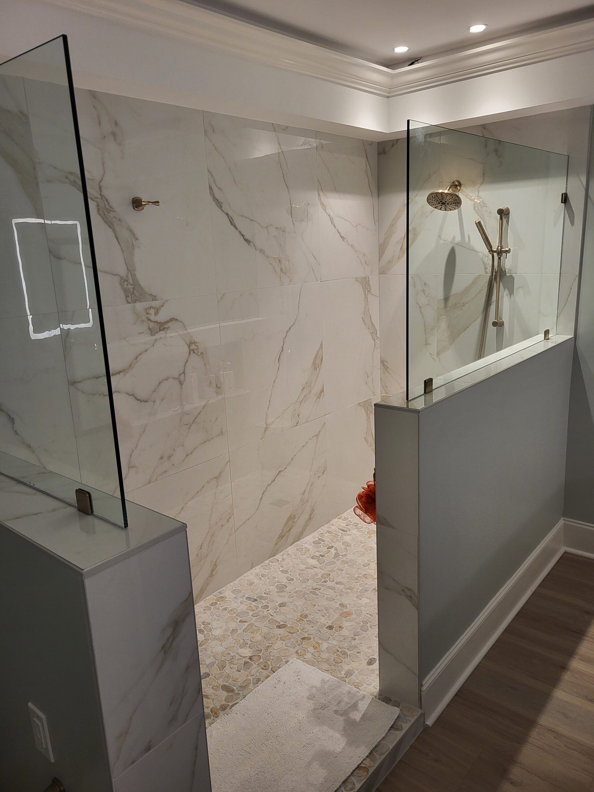A modern walk-in shower with marble-patterned walls, glass panels, and a pebble floor. It has gold fixtures and a gray and white base.