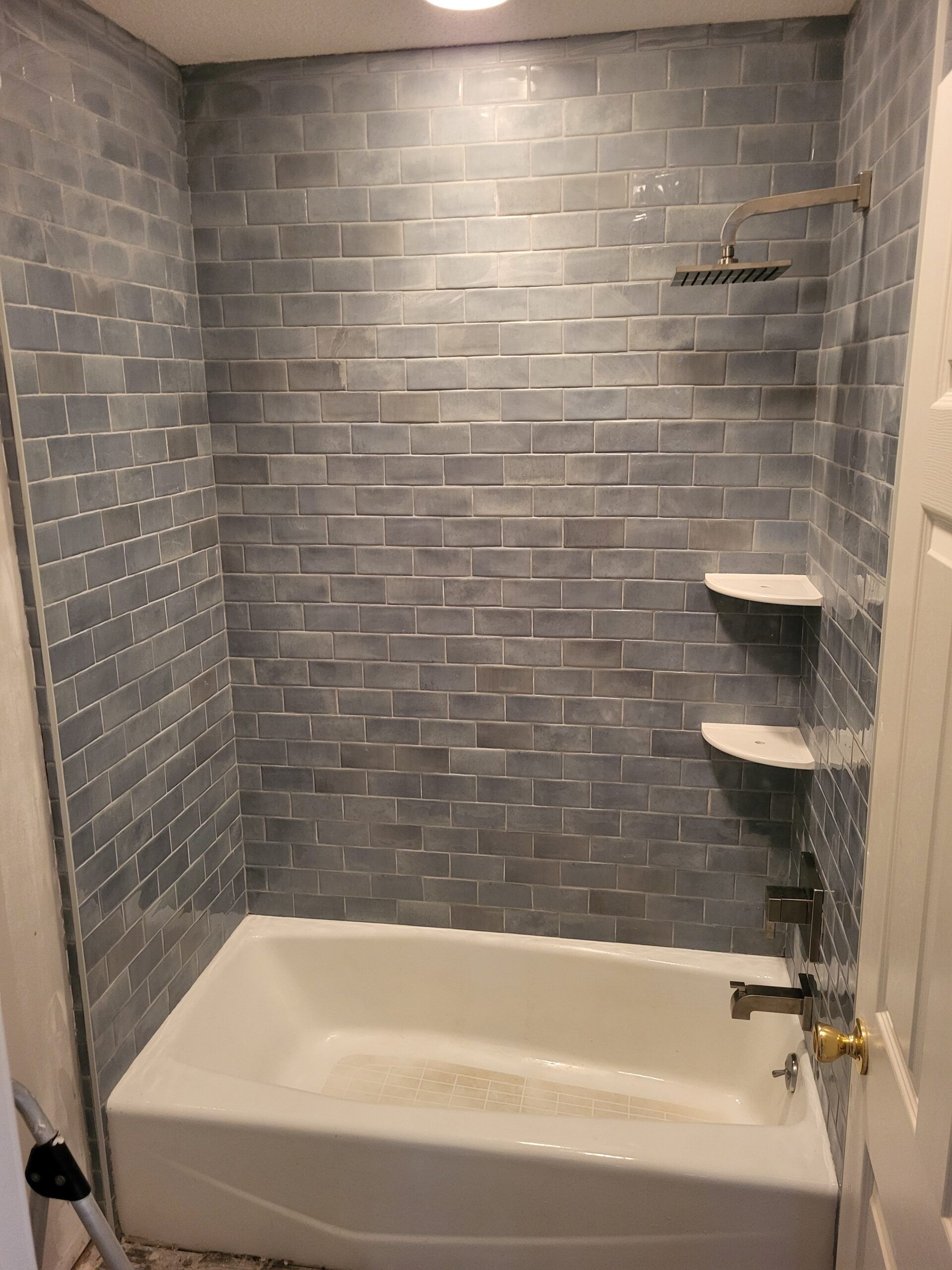 A newly tiled gray shower with a white bathtub, shelves, and bronze fixtures. The setting is a bathroom.