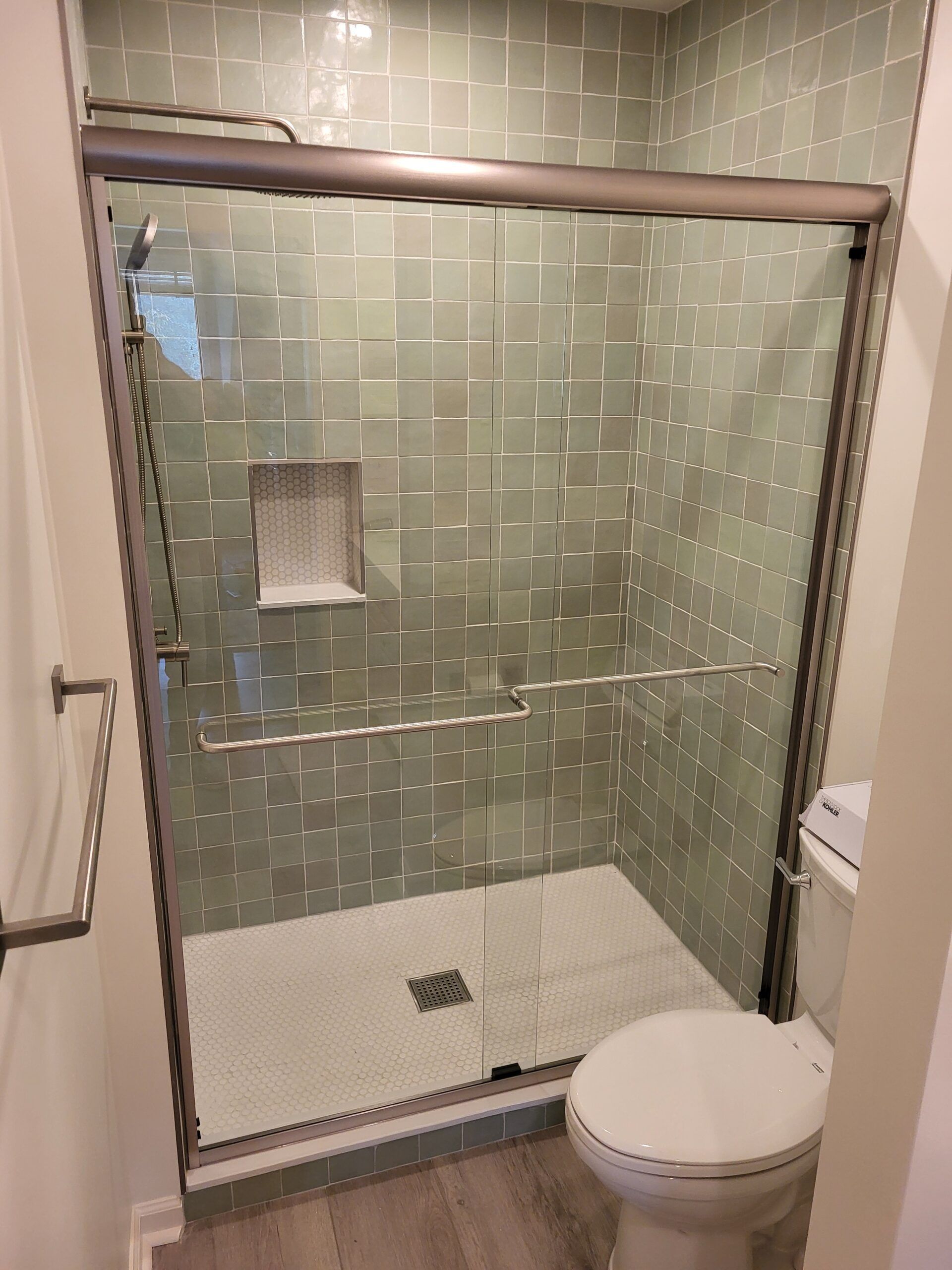 A small bathroom with a glass-enclosed shower, toilet, and towel rack. The shower has light green tiled walls and a white shower base.