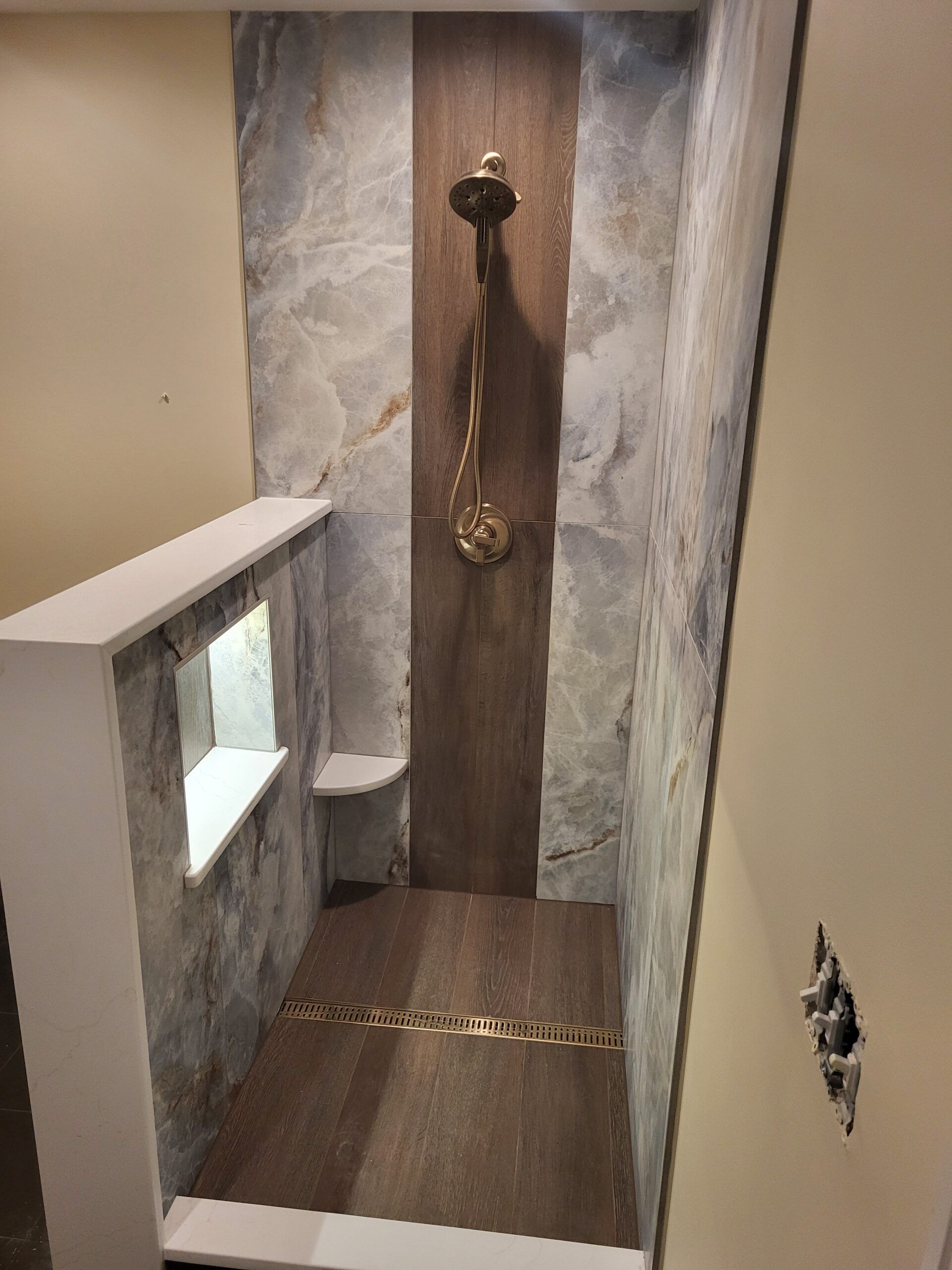 Shower with stone tile walls, wooden accent panel, gold shower head, and brown tile floor. White ledge with window and small seat.