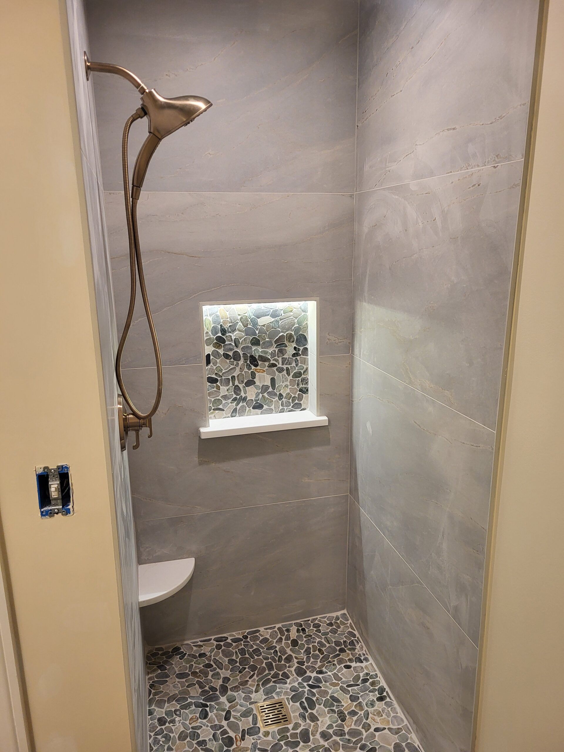 Small, modern shower stall with gray walls and pebble-style floor. A bronze showerhead and a recessed window with decorative tile are visible.