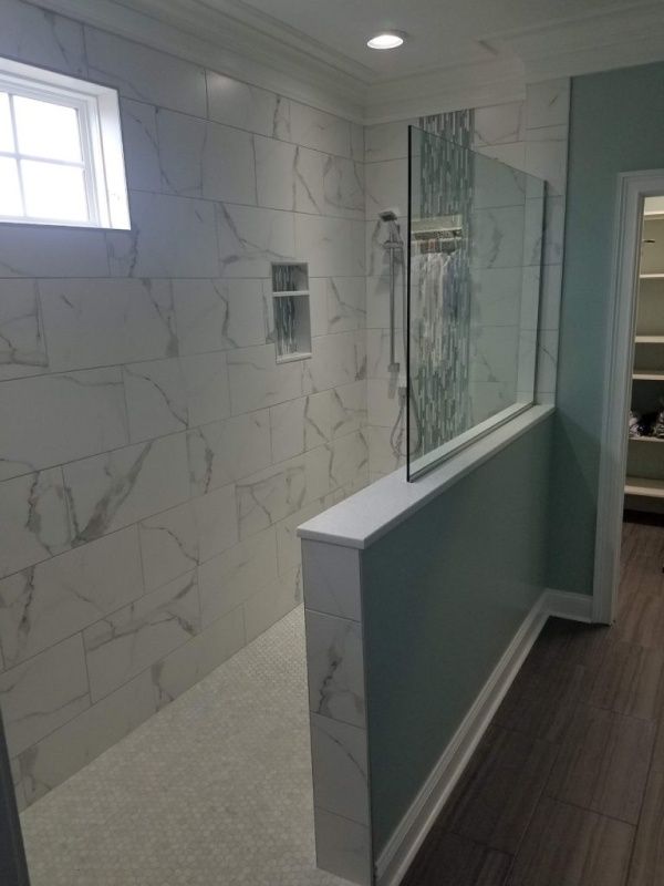 A modern, tiled bathroom with a walk-in shower. White marble-look tiles line the walls and floor. A partial glass shower wall sits on a teal-painted knee wall.