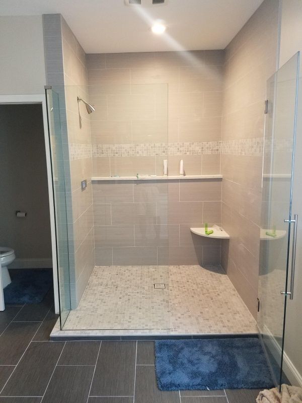 Spacious, accessible shower with gray tile walls, a pebble floor, and glass doors. Includes a built-in shelf and a corner seat.