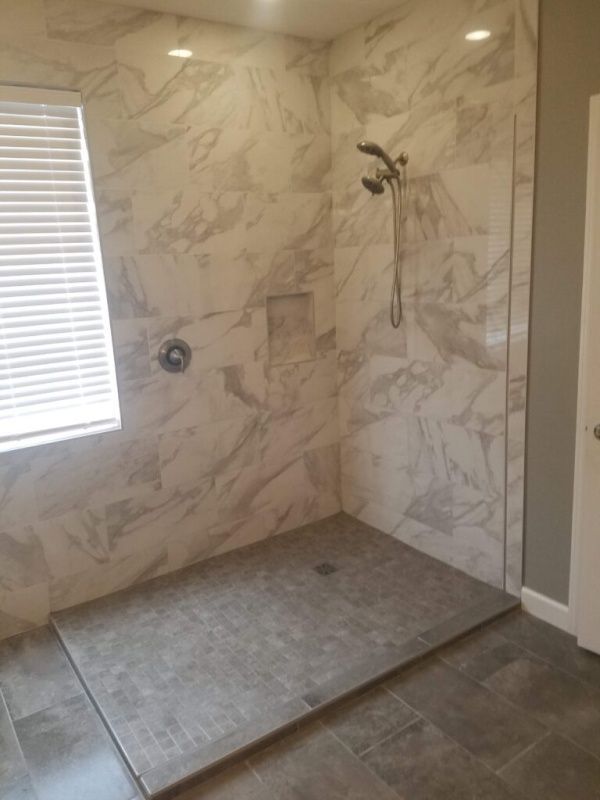 A modern bathroom with a large shower featuring gray tile floor and marble-like patterned walls. A window with blinds is visible.