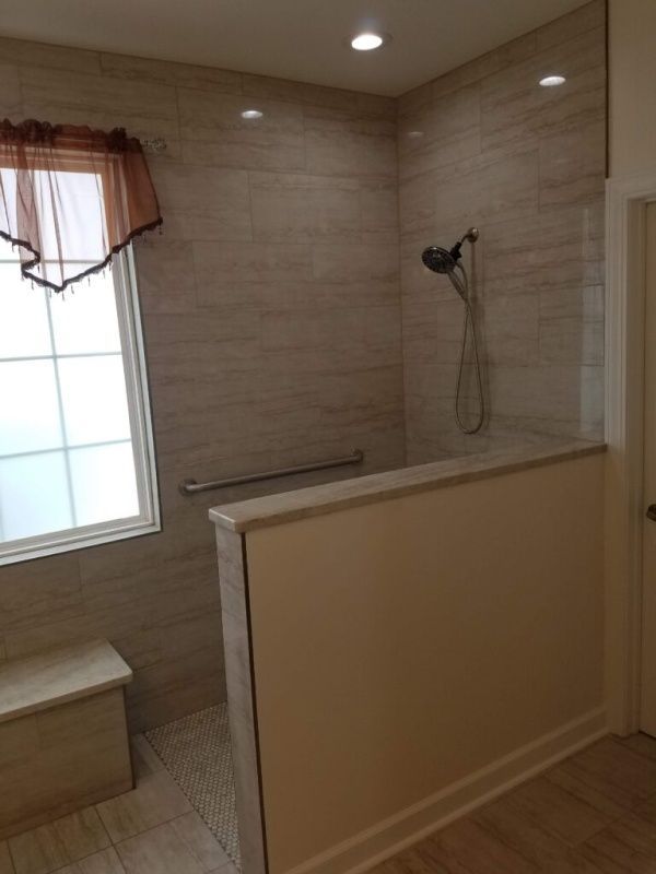 Accessible shower with tan tile walls, a glass partition, and a wall-mounted handrail. There's a window with a bench and a decorative curtain.