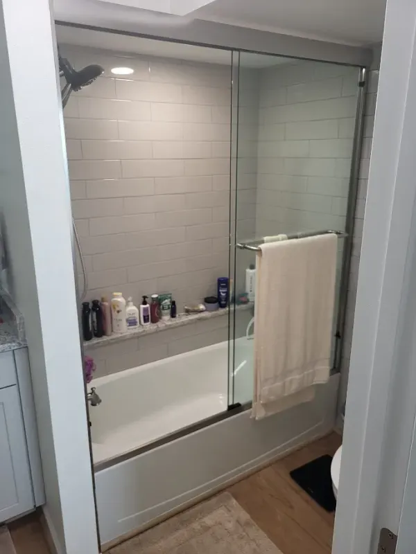 A white bathtub with a glass sliding door, built-in shelf with toiletries, and a towel hanging on the door.