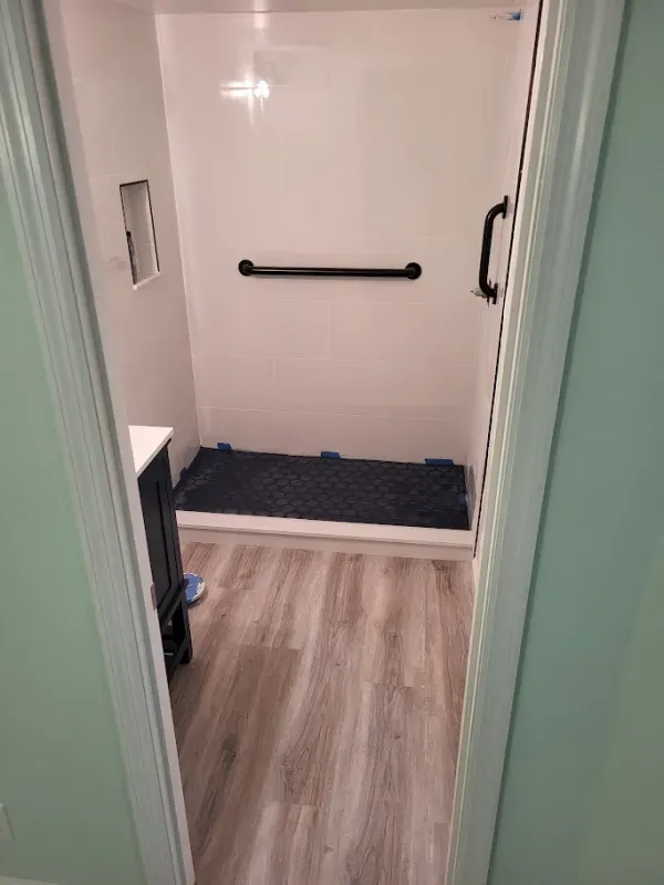 A bathroom with light wood-look flooring and a white shower with a black grab bar. A dark blue mat is in the shower.