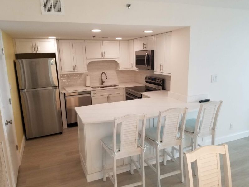 A bright white kitchen with a stainless steel refrigerator, dishwasher, and bar seating.