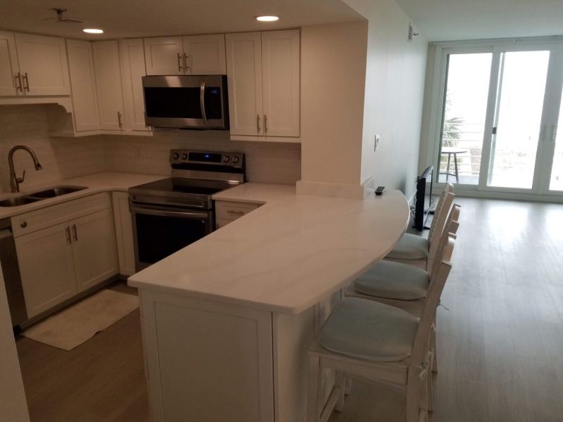 Bright kitchen with white cabinets and countertops. A stainless steel microwave and stove are visible. Bar seating faces the kitchen island.