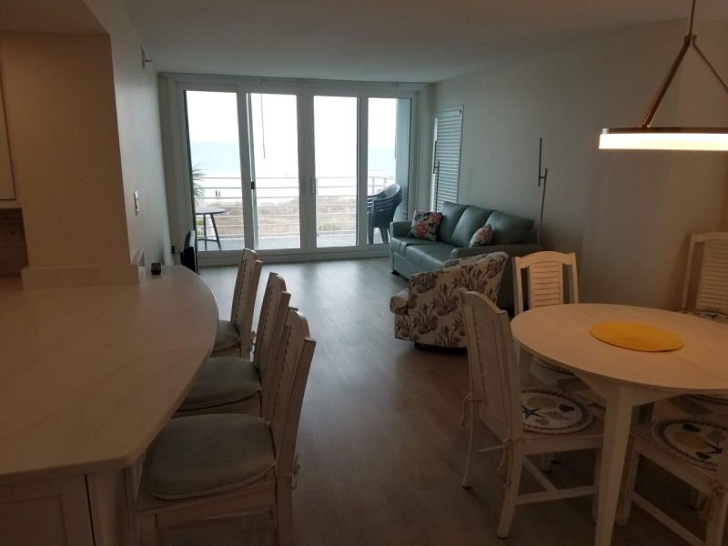 Living room with ocean view, light wood floors, and white furniture. Features a blue sofa, dining table, and bar seating.