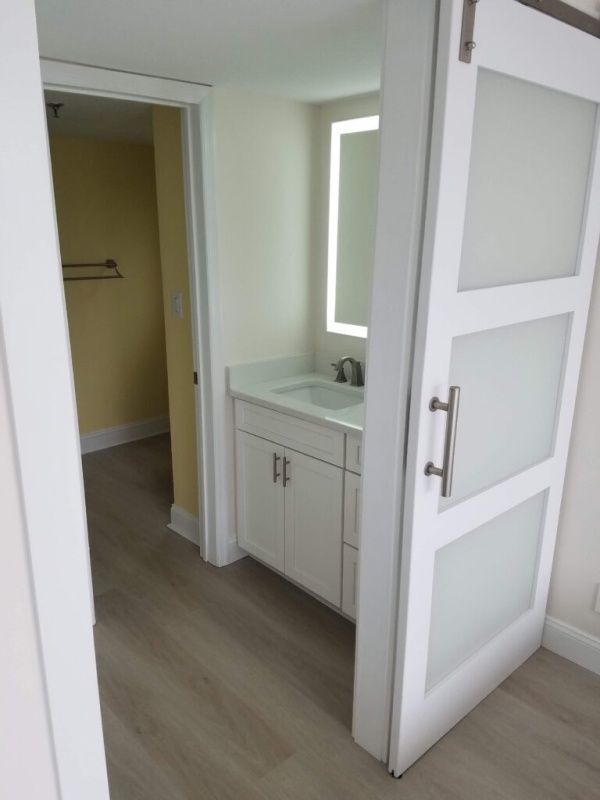 Bathroom with sliding frosted glass door, white vanity, and lighted mirror. Light gray flooring and pale yellow doorway visible.