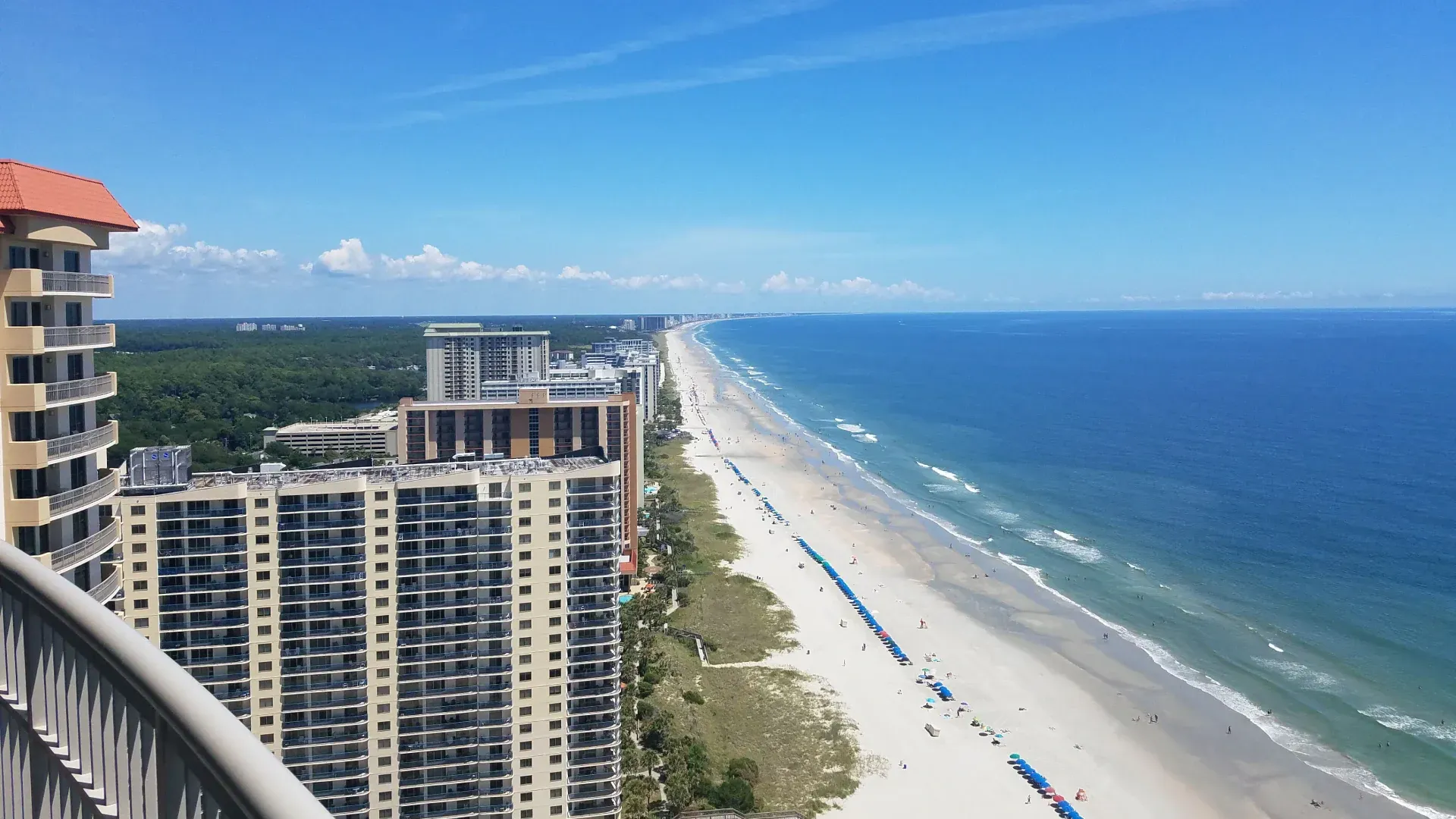 Oceanfront view from a high-rise building, showing a sandy beach, blue ocean, and clear sky. Several buildings are in the frame.