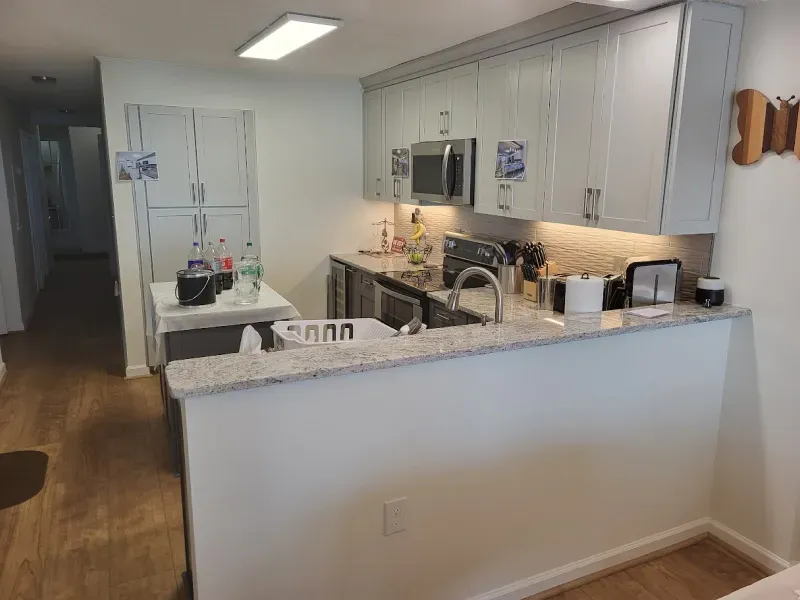 A small kitchen with light gray cabinets and countertops. A sink area, microwave, and breakfast bar are visible.