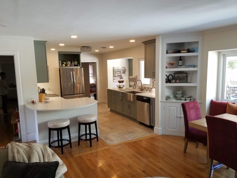 Open-concept kitchen with a breakfast bar, stainless steel appliances, and built-in shelving. Light wood floors and gray cabinets contrast with white countertops.