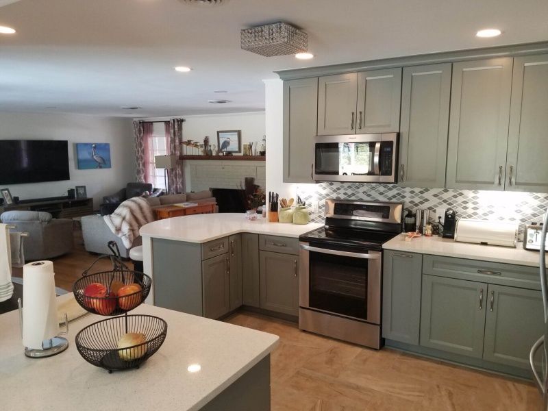 A modern kitchen with light blue cabinets, white countertops, stainless steel appliances, and a view into the living room.