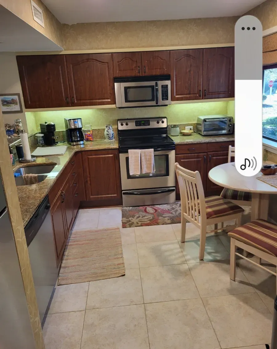 A kitchen with brown cabinets, stainless steel appliances, and a small round table. Beige tile flooring and a light-colored rug are present.