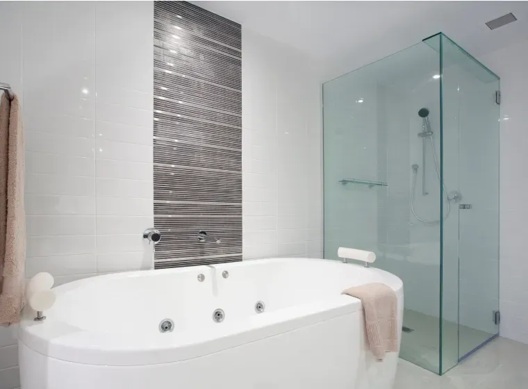 Modern bathroom with a white oval jacuzzi tub and a glass shower. A gray tiled wall provides a contrasting backdrop.