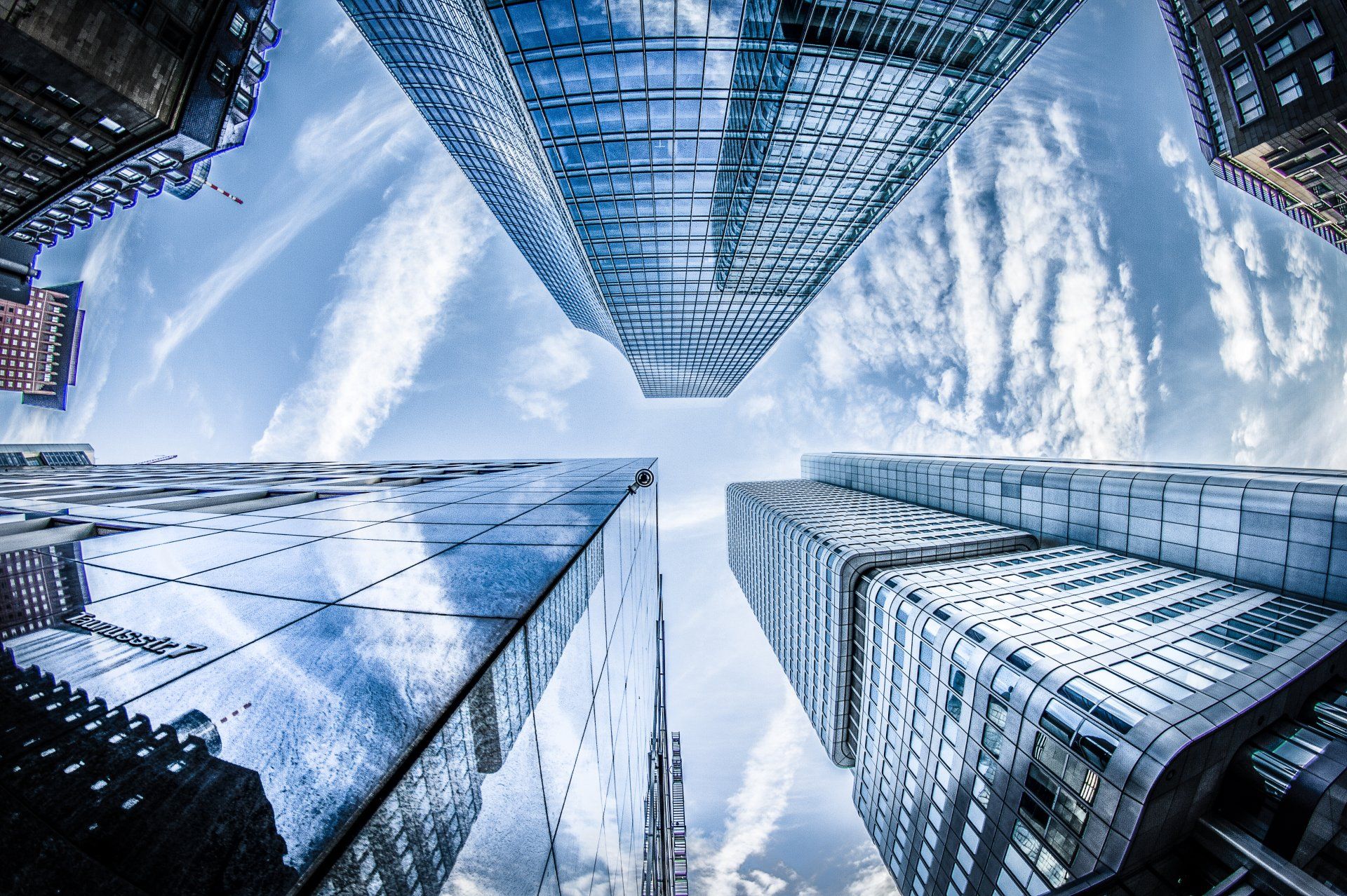 Looking up at tall, modern skyscrapers with reflective glass facades against a blue sky, some cloud trails.