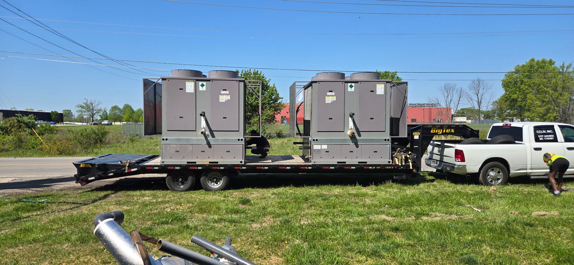 Two gray utility trailers with equipment parked on grass beside a white pickup truck under a blue sky