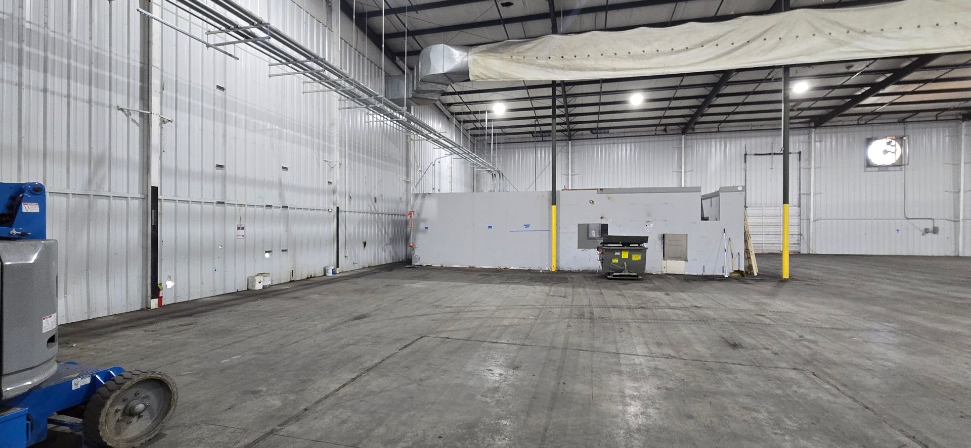 Empty industrial warehouse with concrete floor, white walls, overhead lights, and a forklift near the back wall