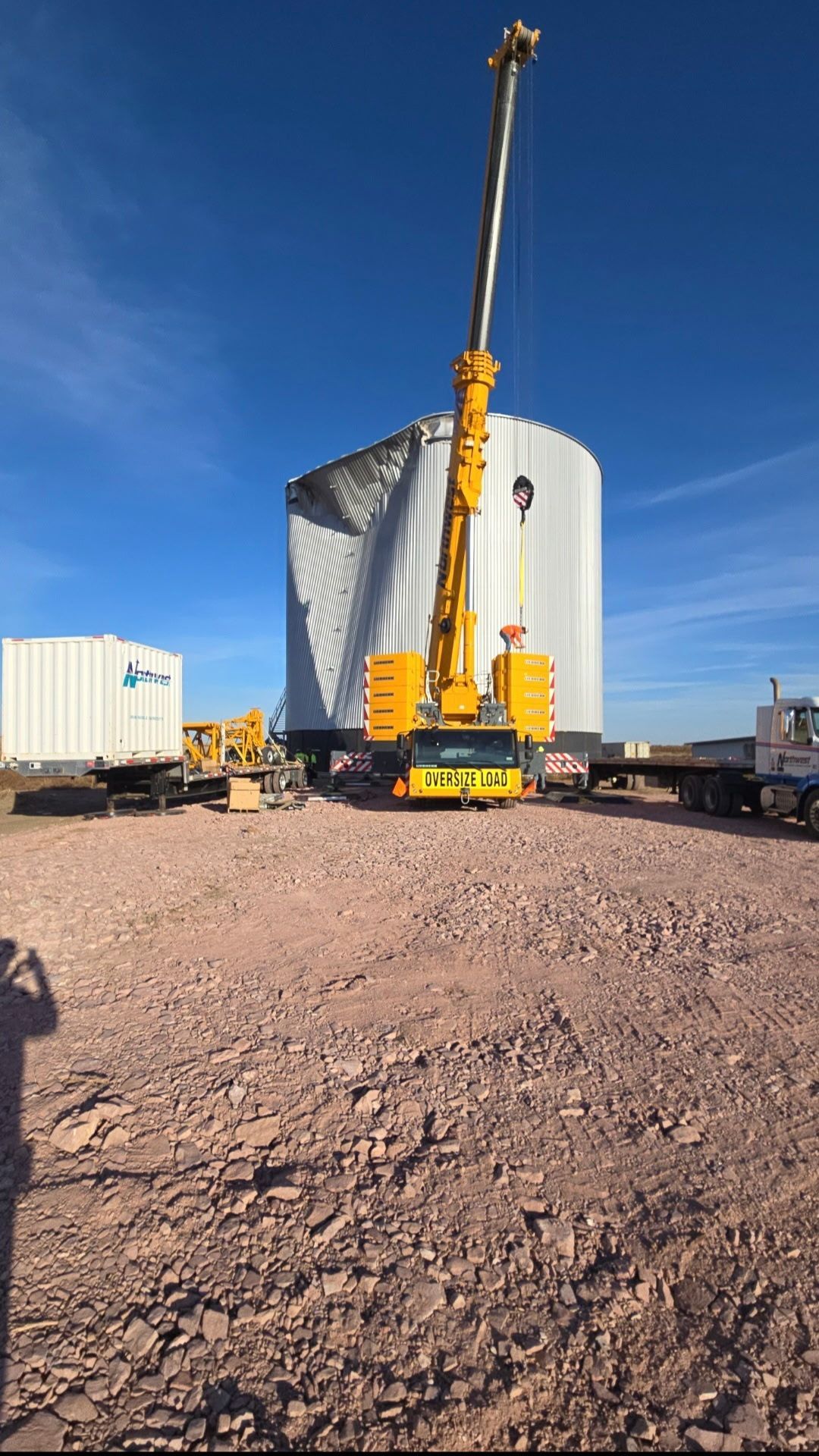 A yellow mobile crane stands on a rocky site next to a large, metallic industrial tank with a dented top section.