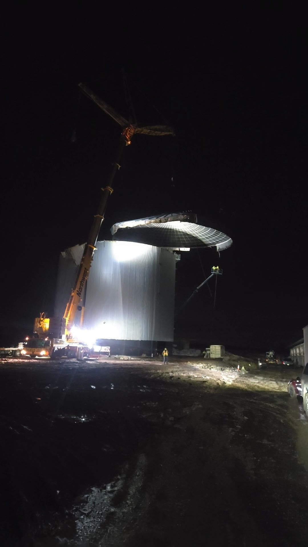 A crane lifts a large, circular metal roof onto a tall industrial storage tank at night under floodlights.
