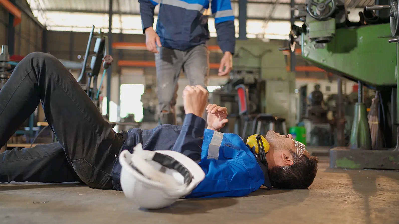 A man is laying on the floor in a factory with a hard hat on his head.
