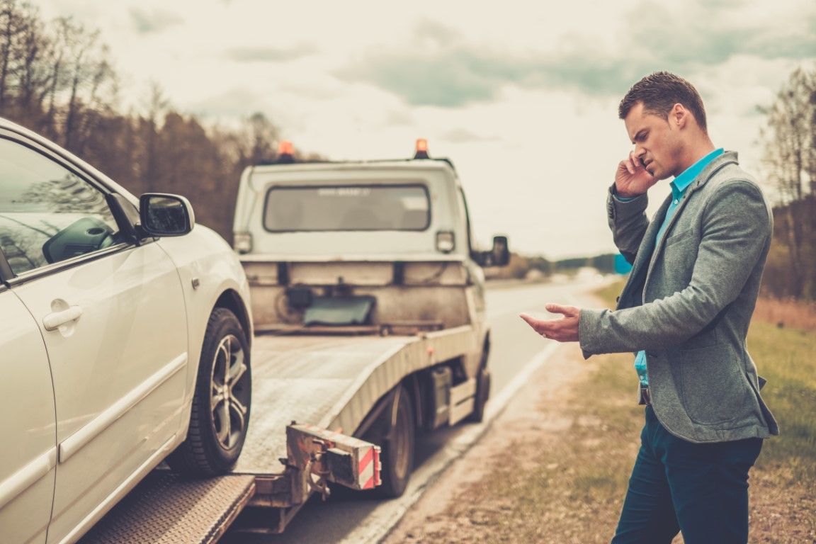 A man stands by a tow truck on the roadside, talking on his phone while his white car is being loaded onto the flatbed.