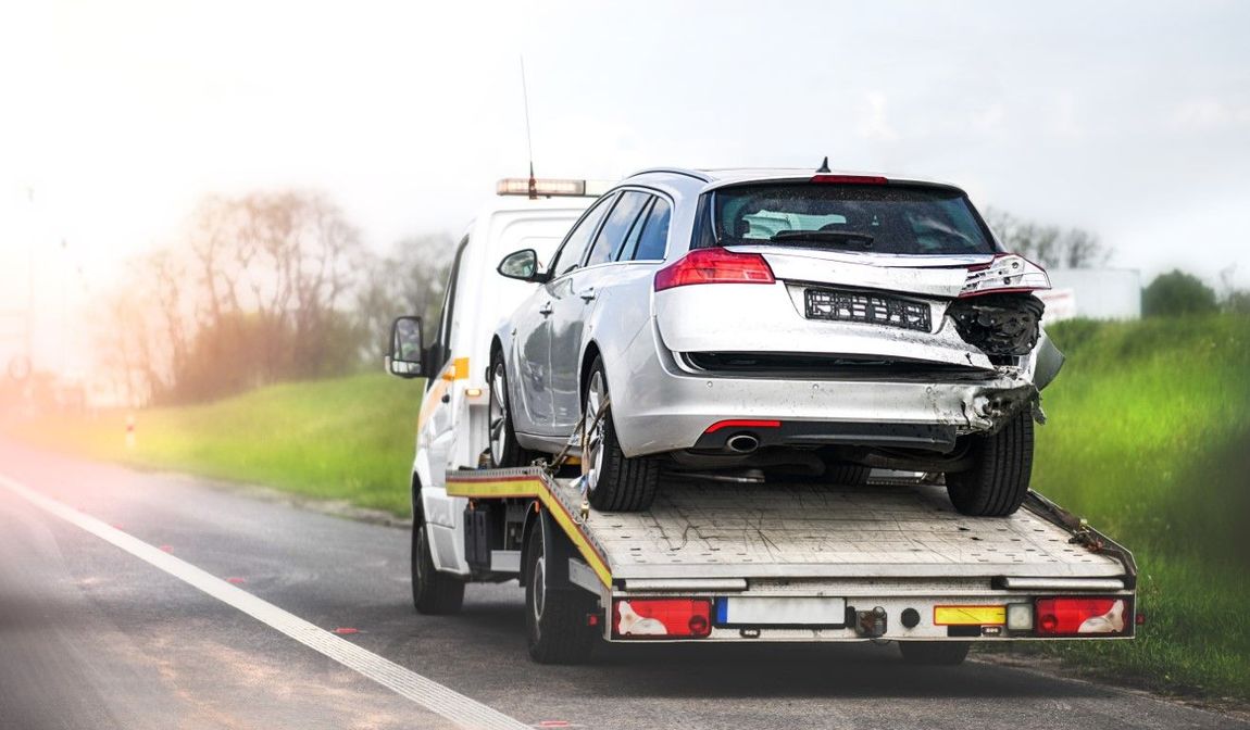 A tow truck transports a silver car with rear-end damage on a highway, likely after an accident or breakdown.