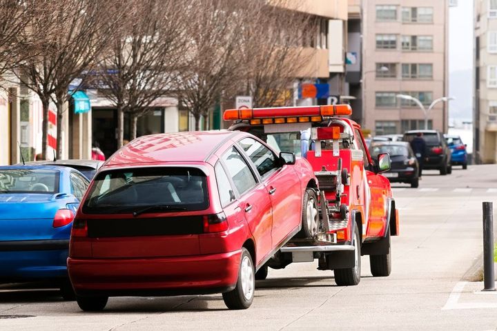 A red tow truck hauls a red car by its front wheels in a city street, possibly due to a parking violation or breakdown.
