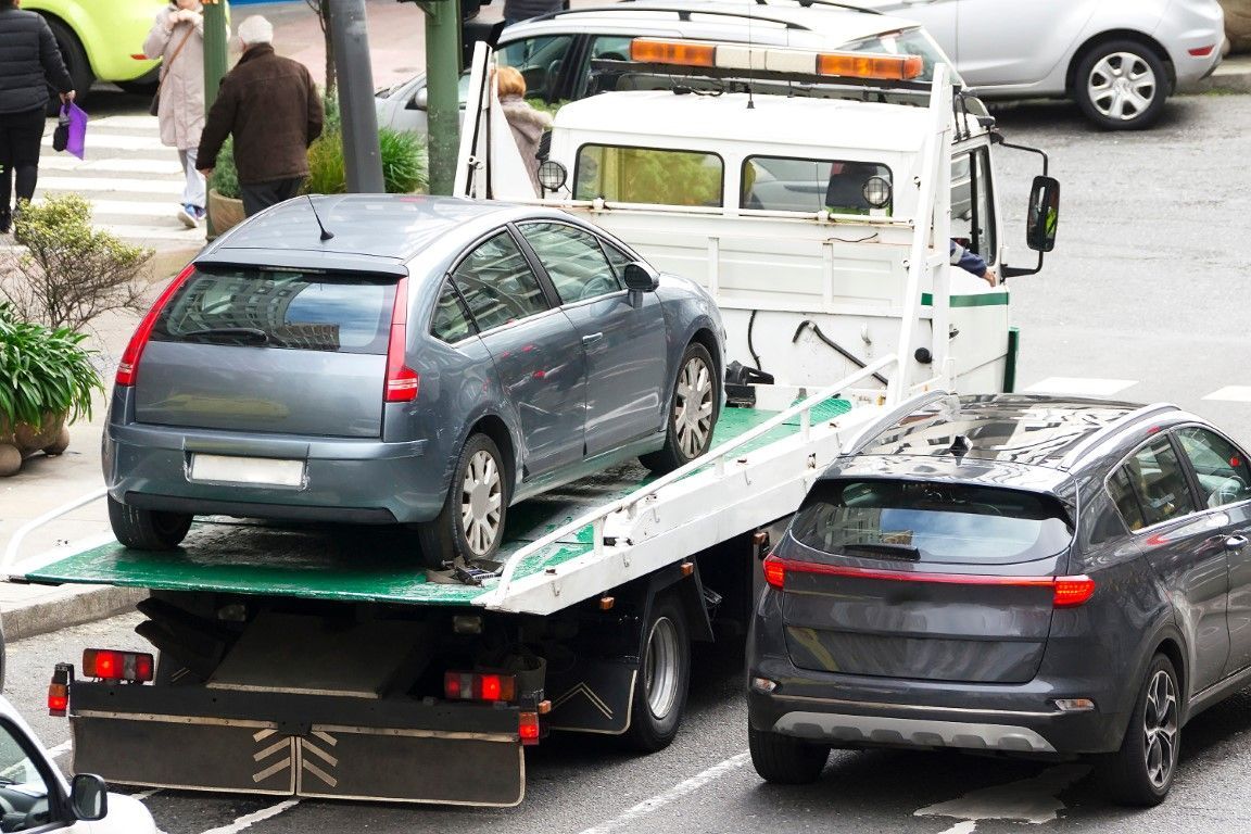 Gray hatchback car loaded onto a flatbed tow truck in a busy urban area with pedestrians and traffic nearby.